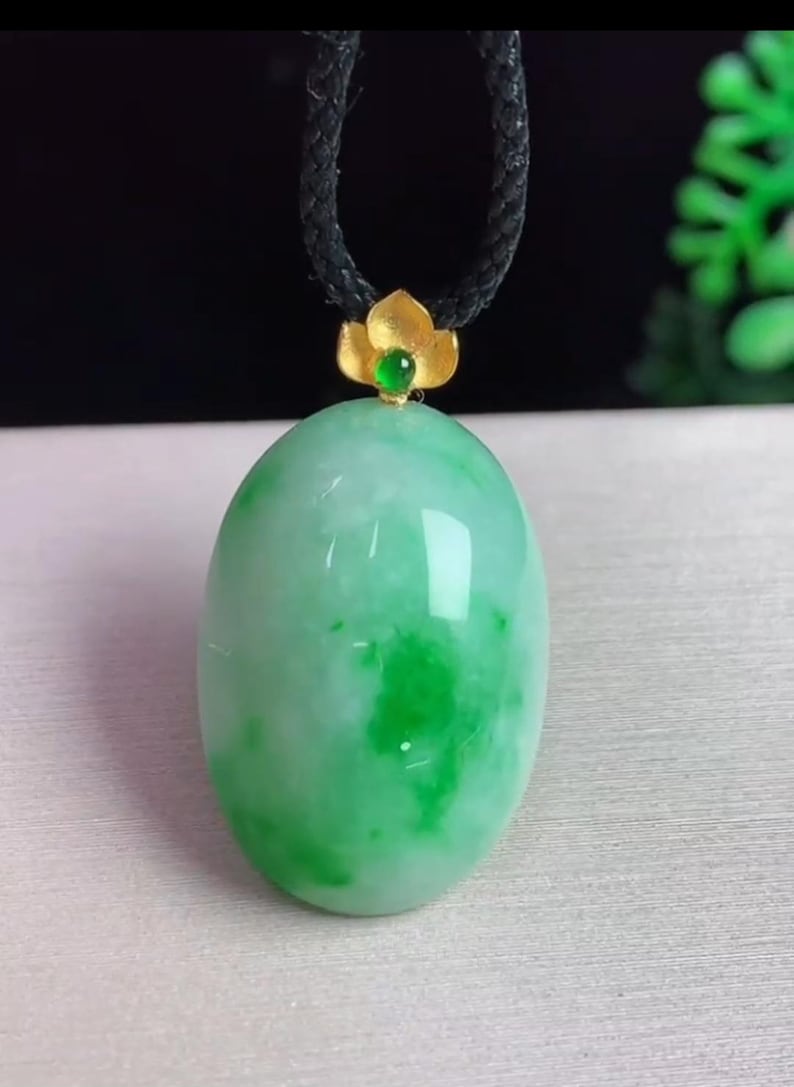 May include: A jade pendant necklace featuring a large, oval-shaped, light green jade stone. The jade has darker green marbling. The pendant is topped with a gold-colored flower and a small green gemstone, suspended from a black cord.