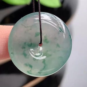 Puede incluir: Colgante redondo de jade transl&uacute;cido con un patr&oacute;n moteado verde y blanco. El colgante est&aacute; suspendido por un cord&oacute;n marr&oacute;n oscuro. El jade tiene un acabado pulido y brillante.