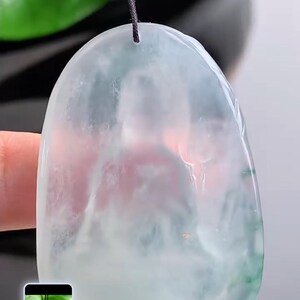 May include: A translucent jade pendant, with a light green and white colour scheme, suspended by a black cord. The jade has a smooth, polished surface and an irregular shape. The background is blurred.