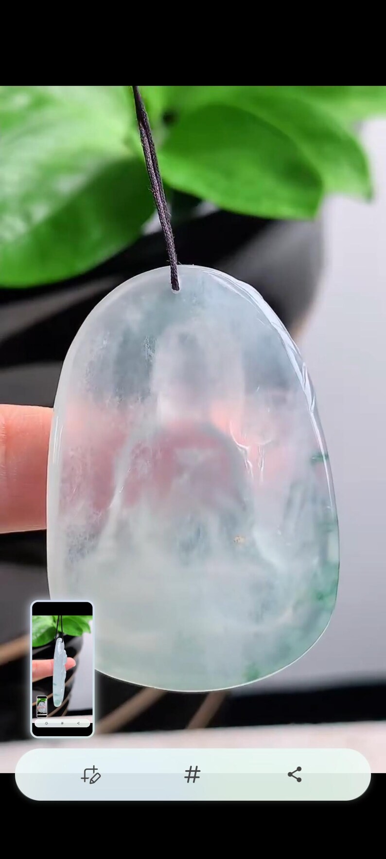 May include: A translucent jade pendant, with a light green and white colour scheme, suspended by a black cord. The jade has a smooth, polished surface and an irregular shape. The background is blurred.