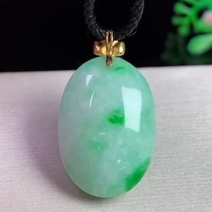 May include: Oval jade pendant with a gold-colored bail, suspended from a black cord. The jade is a mix of light green and white, with darker green veining. The pendant has a polished, glossy finish.