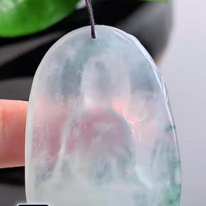 May include: A translucent jade pendant with a rounded top and a dark cord. The pendant has a light green and white colour scheme. The background includes green leaves and a blurred view of a hand holding the pendant.
