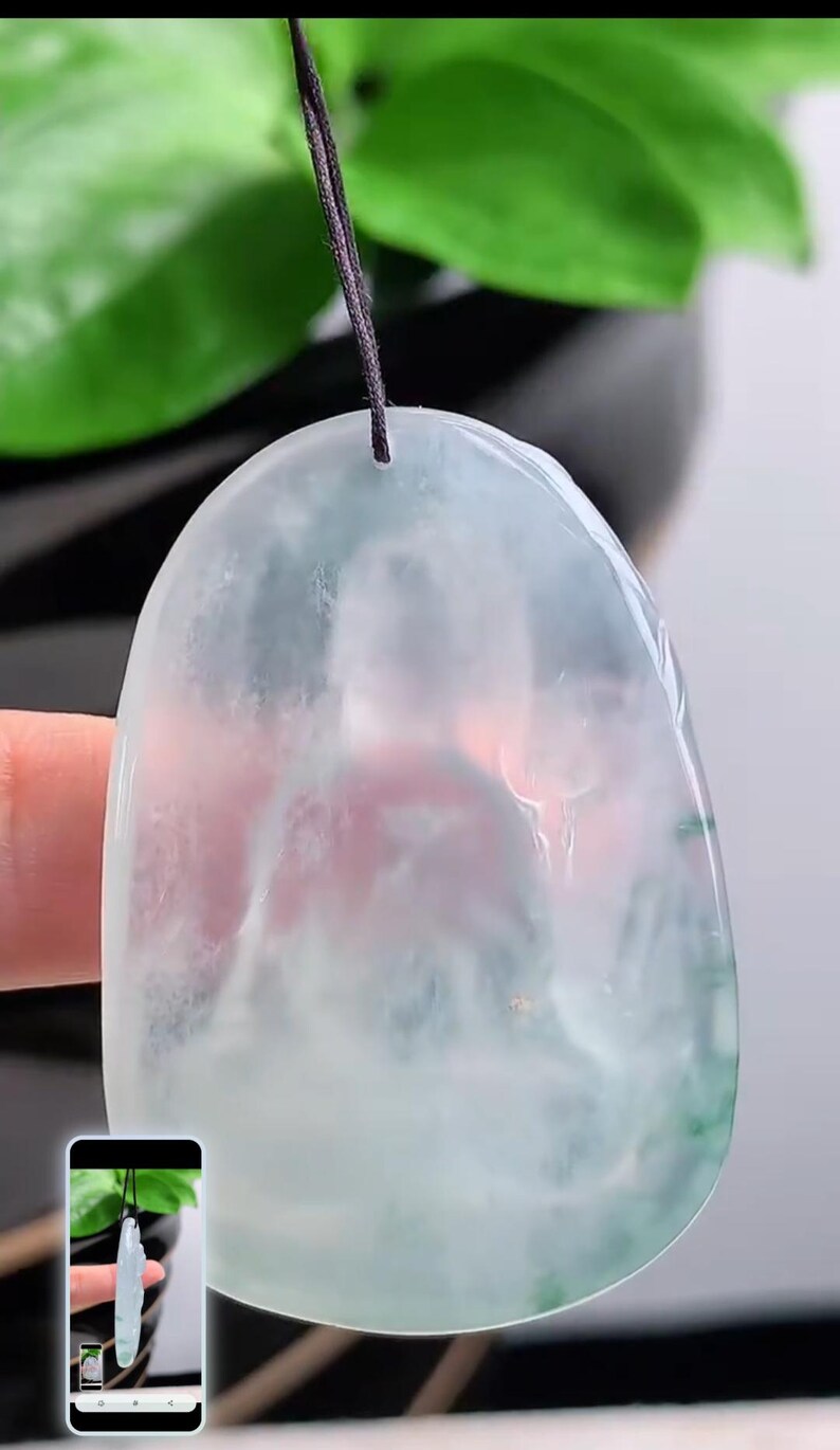 May include: A translucent jade pendant with a rounded top and a dark cord. The pendant has a light green and white colour scheme. The background includes green leaves and a blurred view of a hand holding the pendant.
