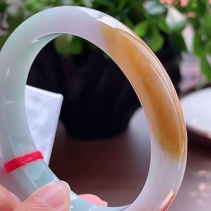 Può includere: Un bracciale rigido in giada liscia e lucida, caratterizzato da una base bianca traslucida con macchie verde pallido e una distinta sezione color marrone dorato. Un sottile filo rosso è legato attorno a un lato dell'anello in pietra.