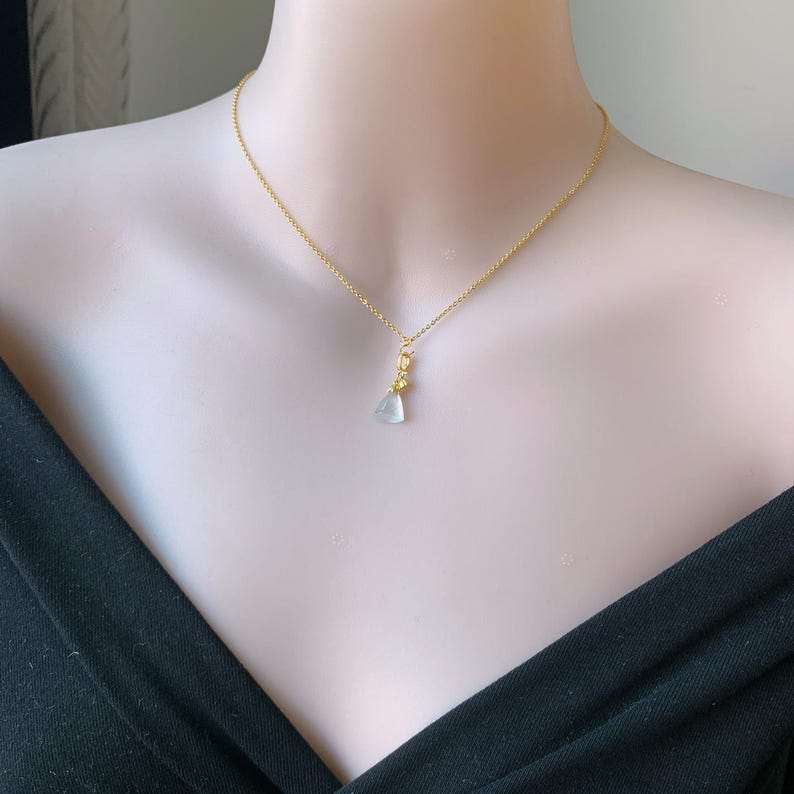 May include: A delicate gold chain necklace featuring a small, translucent, pale green triangular pendant. The pendant is suspended from a gold-toned bail, creating a simple and refined piece of jewelry that rests against the collarbone area.