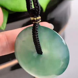 Pu&ograve; includere: Un ciondolo di giada verde a forma di cerchio con un cordoncino nero attaccato. Il ciondolo &egrave; appeso a un cordoncino nero.