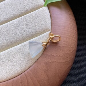 May include: A triangular pendant featuring a translucent, pale grey stone set in a polished gold-tone metal cap. The cap includes a small, rounded bail at the top for attachment, showcasing a smooth, minimalist design with clean, geometric lines.