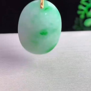 May include: A light green oval pendant with darker green spots, suspended by a gold-colored bail and a black cord. The pendant has a smooth, polished surface and is set against a neutral background.