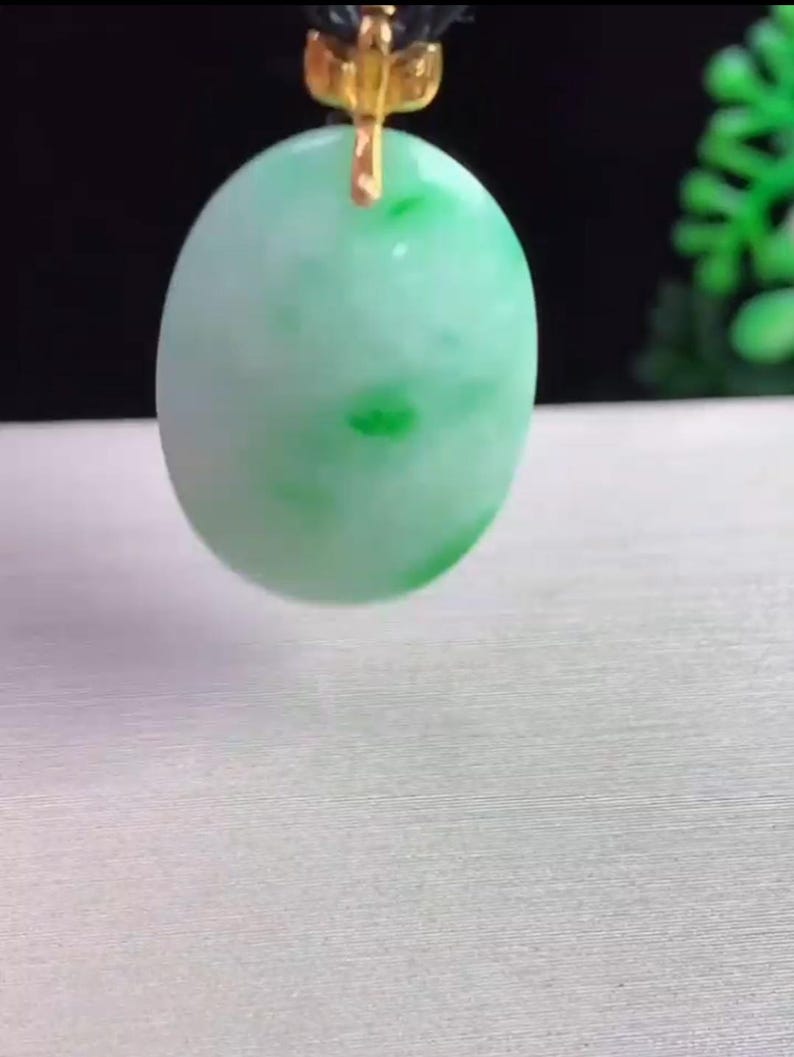 May include: A light green oval pendant with darker green spots, suspended by a gold-colored bail and a black cord. The pendant has a smooth, polished surface and is set against a neutral background.