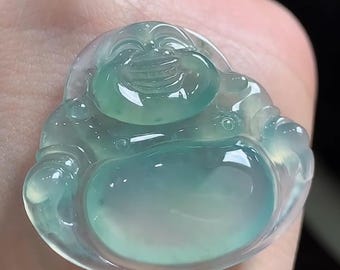 May include: A translucent, light green jade carving of a smiling Buddha figure. The stone has a smooth, polished surface with a glossy finish, showcasing natural variations in colour and clarity throughout the rounded, seated form of the pendant.