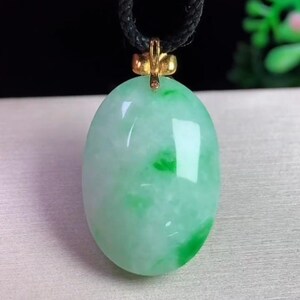 May include: Oval jade pendant with a gold-colored bail, suspended from a black cord. The jade is a mix of light green and white, with darker green patches. The pendant is smooth and polished, reflecting light.