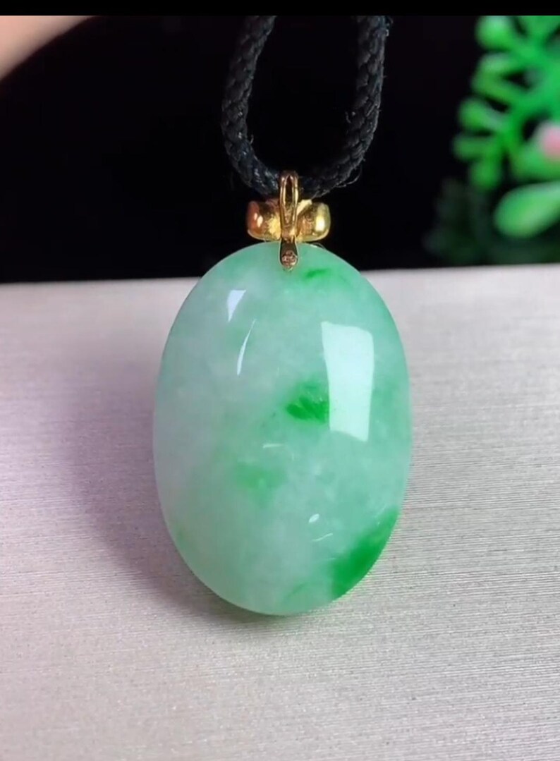 May include: Oval jade pendant with a gold-colored bail, suspended from a black cord. The jade is a mix of light green and white, with darker green patches. The pendant is smooth and polished, reflecting light.