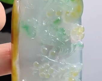 May include: A jade pendant with a floral design. The pendant is a light green and yellow color, with a dark yellow bead at the top. The pendant is held by a black cord.