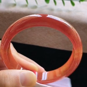 Può includere: Un bracciale rigido rosso traslucido, liscio e lucido, con un profilo arrotondato. Il materiale presenta una finitura brillante che riflette la luce, mostrando variazioni nell'intensità del colore lungo tutta la struttura circolare.