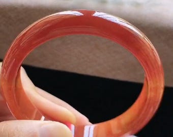 Puede incluir: Una pulsera tipo brazalete de color rojo translúcido, lisa y pulida, con un perfil redondeado. El material presenta un acabado brillante que refleja la luz, mostrando variaciones en la intensidad del color en toda la estructura circular.