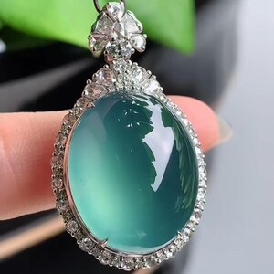 May include: A jade pendant with a green gemstone surrounded by a halo of small diamonds. The pendant is attached to a black cord.