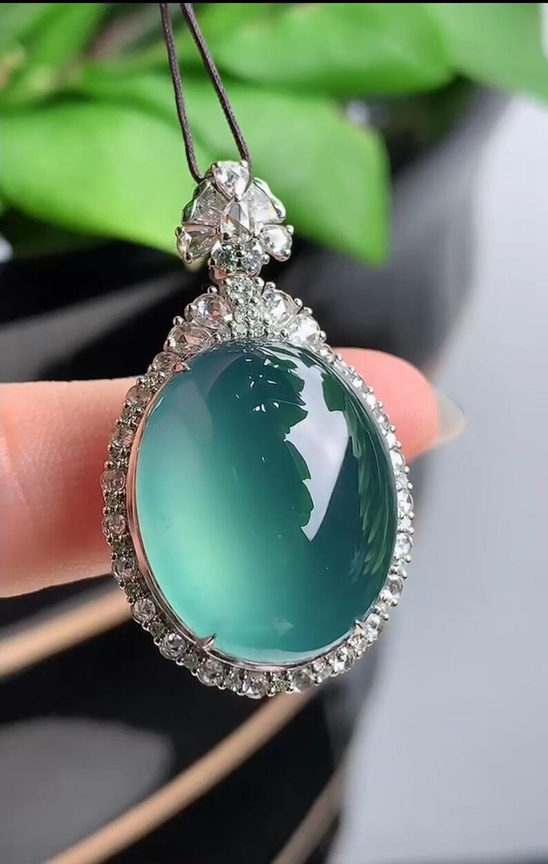 May include: A jade pendant with a green gemstone surrounded by a halo of small diamonds. The pendant is attached to a black cord.