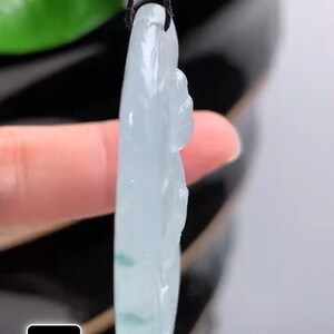 May include: A translucent jade pendant carved in the shape of a seated Buddha. The pendant is light green with darker green inclusions and is suspended from a black cord. The pendant is held up by a finger.