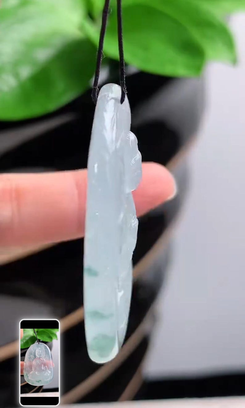 May include: A translucent jade pendant carved in the shape of a seated Buddha. The pendant is light green with darker green inclusions and is suspended from a black cord. The pendant is held up by a finger.