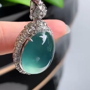 May include: A silver pendant with a large, oval-shaped, light blue gemstone surrounded by small, clear gemstones. The pendant is hanging from a black cord.