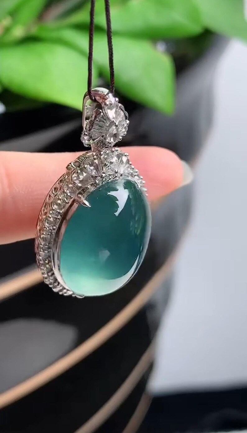 May include: A silver pendant with a large, oval-shaped, light blue gemstone surrounded by small, clear gemstones. The pendant is hanging from a black cord.