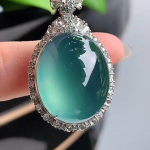 May include: A jade pendant with a green oval stone surrounded by a halo of small diamonds. The pendant is attached to a black cord.