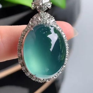 May include: A jade pendant with a round, light green jade stone surrounded by a halo of small diamonds. The pendant is attached to a black cord.