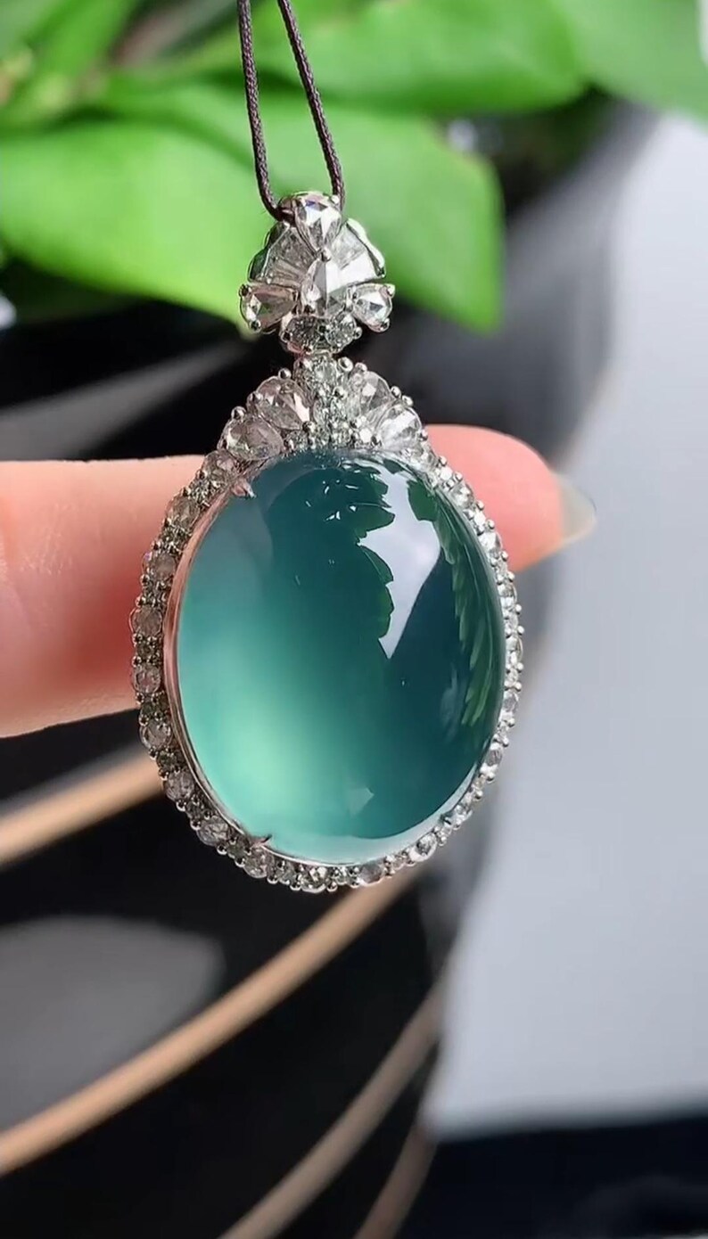 May include: A jade pendant with a round, light green jade stone surrounded by a halo of small diamonds. The pendant is attached to a black cord.