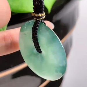 Pu&ograve; includere: Un ciondolo di giada di colore verde chiaro e forma ovale liscia. Il ciondolo &egrave; attaccato a un cordino nero con accenti dorati.