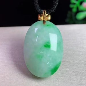 May include: Oval jade pendant with a gold-colored bail, suspended from a black cord. The jade is a mix of light green and white, with darker green patches. The pendant has a polished, glossy finish.
