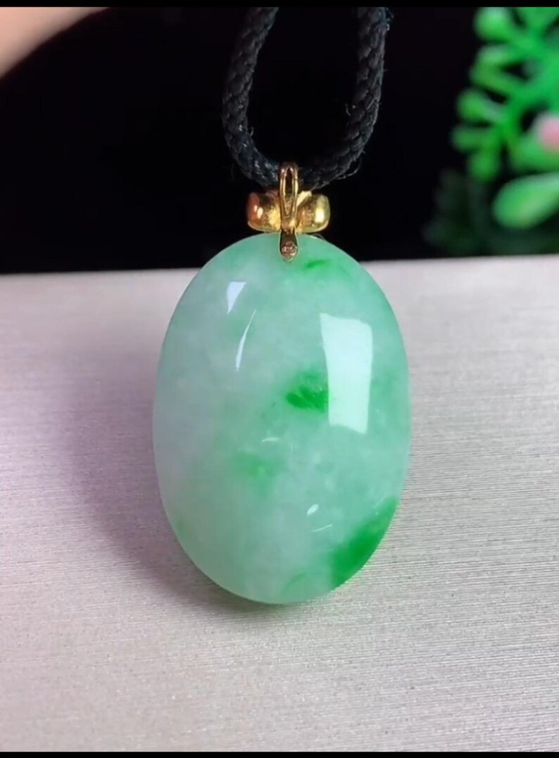 May include: Oval jade pendant with a gold-colored bail, suspended from a black cord. The jade is a mix of light green and white, with darker green patches. The pendant has a polished, glossy finish.