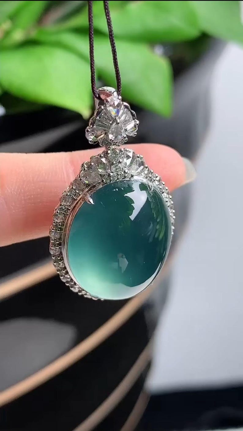 May include: A jade pendant necklace with a round, green jade stone set in a silver frame with a border of small diamonds.