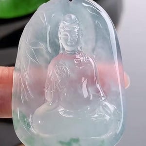 May include: A translucent jade pendant featuring a seated Buddha figure. The pendant is oval-shaped with a carved design of the Buddha in a meditative pose, with bamboo leaves on the sides. The jade has shades of green and white.