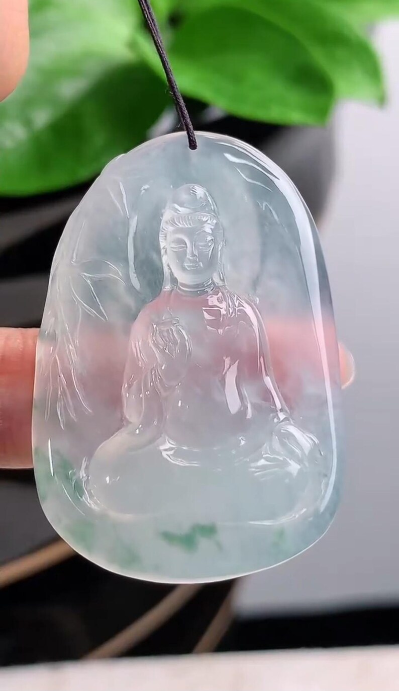 May include: A translucent jade pendant featuring a seated Buddha figure. The pendant is oval-shaped with a carved design of the Buddha in a meditative pose, with bamboo leaves on the sides. The jade has shades of green and white.