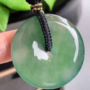 Pu&ograve; includere: Un ciondolo di giada verde su una collana di cordoncino nero. Il ciondolo &egrave; rotondo e ha un foro al centro.