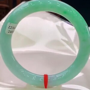 Può includere: Un bracciale rigido in giada verde chiaro, liscio e traslucido, con finitura lucida. Un piccolo adesivo bianco con il testo nero ZZGJ 2907 è attaccato sul lato superiore sinistro e un sottile filo rosso è legato nella parte inferiore.