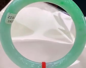 Puede incluir: Un brazalete de jade verde claro, liso y translúcido, con acabado pulido. Una pequeña etiqueta blanca con el texto negro ZZGJ 2907 está adherida en el lado superior izquierdo, y un hilo rojo fino está atado en la sección inferior.