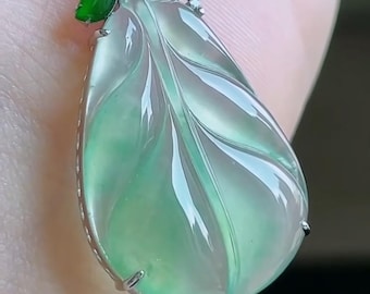 May include: A translucent green jade pendant carved into a leaf shape with a silver-toned metal bail. The pendant features a small, vibrant green jade accent stone and clear, sparkling stones set into the metal top, suspended on a dark cord.