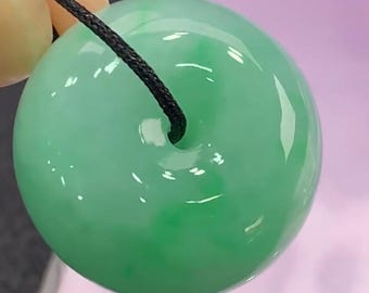 May include: A smooth, circular green jade pendant with a polished, translucent finish and a central hole. A thin black cord passes through the center of the stone, which features mottled light and dark green color variations throughout its surface.
