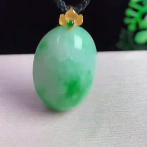 May include: A green jade pendant with a gold-colored floral accent and a black cord. The jade has a smooth, polished surface with varying shades of green. The pendant is oval-shaped and appears to be a necklace.