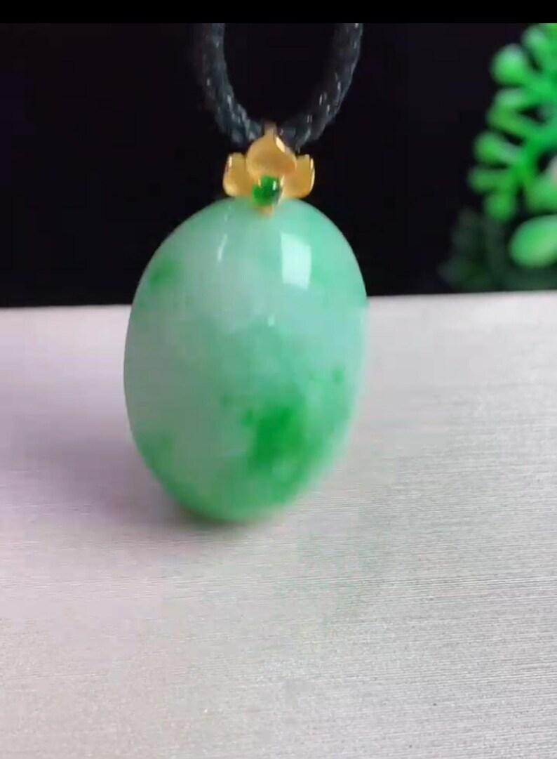 May include: A green jade pendant with a gold-colored floral accent and a black cord. The jade has a smooth, polished surface with varying shades of green. The pendant is oval-shaped and appears to be a necklace.