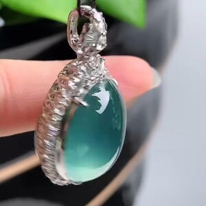 May include: A silver pendant with a light blue gemstone set in a detailed silver frame. The pendant is hanging from a black cord.
