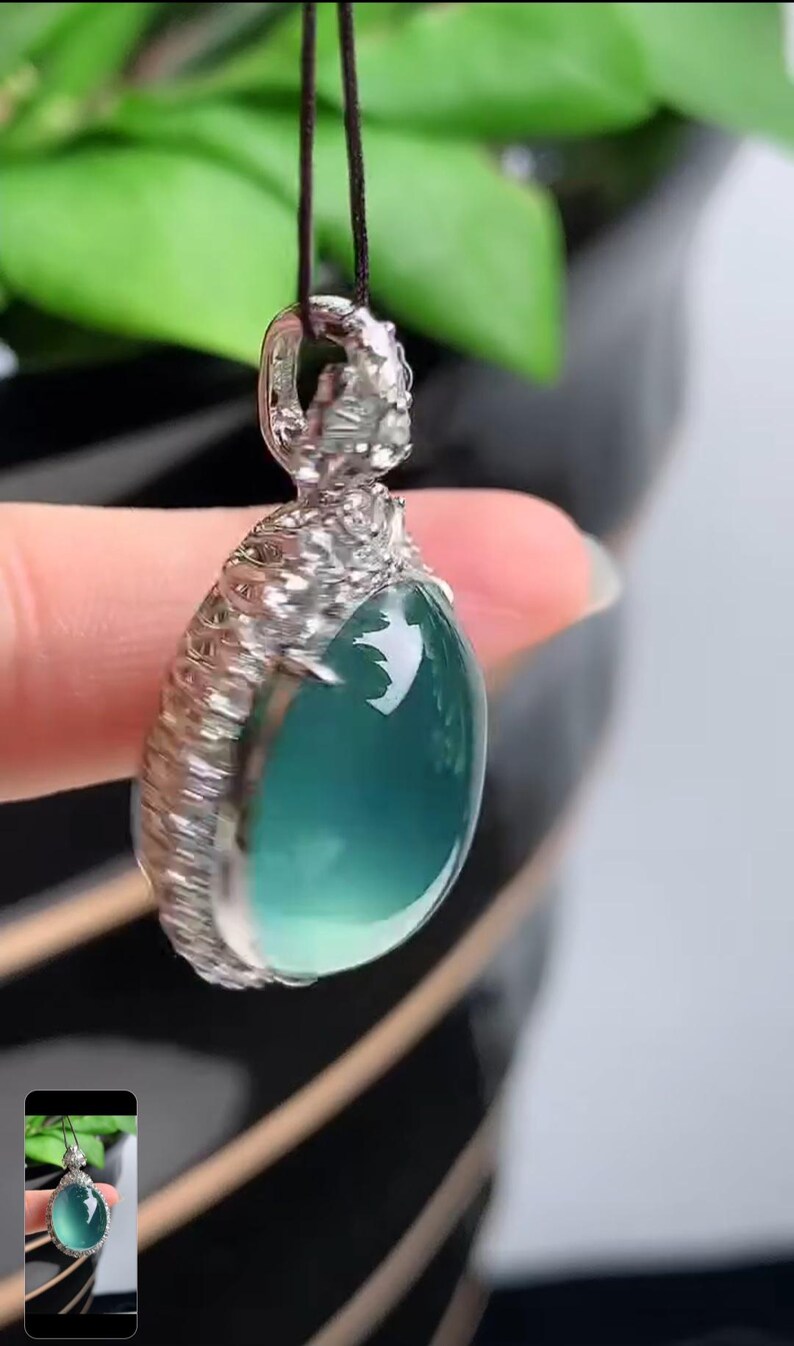 May include: A silver pendant with a light blue gemstone set in a detailed silver frame. The pendant is hanging from a black cord.