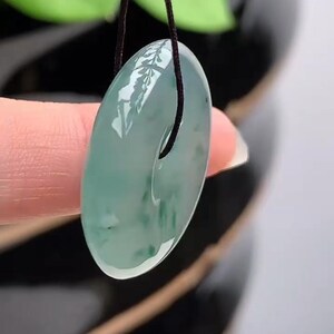 Puede incluir: Un colgante de jade transl&uacute;cido con dise&ntilde;o circular, suspendido de un cord&oacute;n negro. El jade presenta una mezcla de tonos verde claro y oscuro, con un acabado pulido y brillante. El colgante se muestra sobre un fondo borroso de follaje verde.