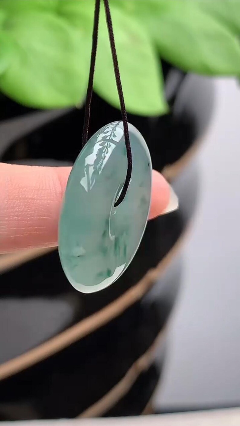 Puede incluir: Un colgante de jade transl&uacute;cido con dise&ntilde;o circular, suspendido de un cord&oacute;n negro. El jade presenta una mezcla de tonos verde claro y oscuro, con un acabado pulido y brillante. El colgante se muestra sobre un fondo borroso de follaje verde.