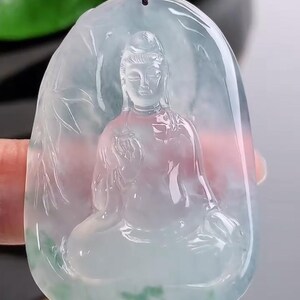 May include: A translucent jade pendant with a carved Buddha figure. The pendant is oval-shaped and features a seated Buddha with intricate details. The jade has a pale green and pink hue, with a black cord for wearing.