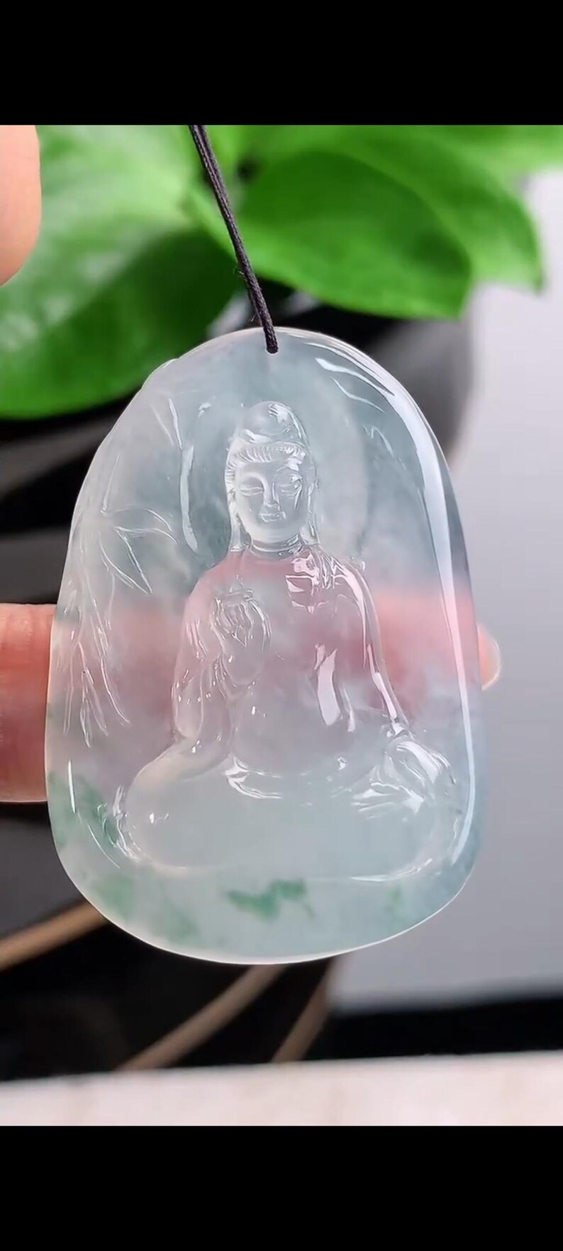 May include: A translucent jade pendant with a carved Buddha figure. The pendant is oval-shaped and features a seated Buddha with intricate details. The jade has a pale green and pink hue, with a black cord for wearing.