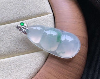 May include: A translucent, pale green jadeite pendant shaped like a gourd, featuring a smooth, polished surface with subtle emerald green inclusions. The pendant is attached to a silver-toned metal bail adorned with a small, round green gemstone.