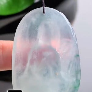 May include: A translucent jade pendant with a light green and pink hue, suspended by a black cord. The smooth, polished surface reflects light, showcasing the natural variations within the stone. The pendant's shape is an elongated oval.