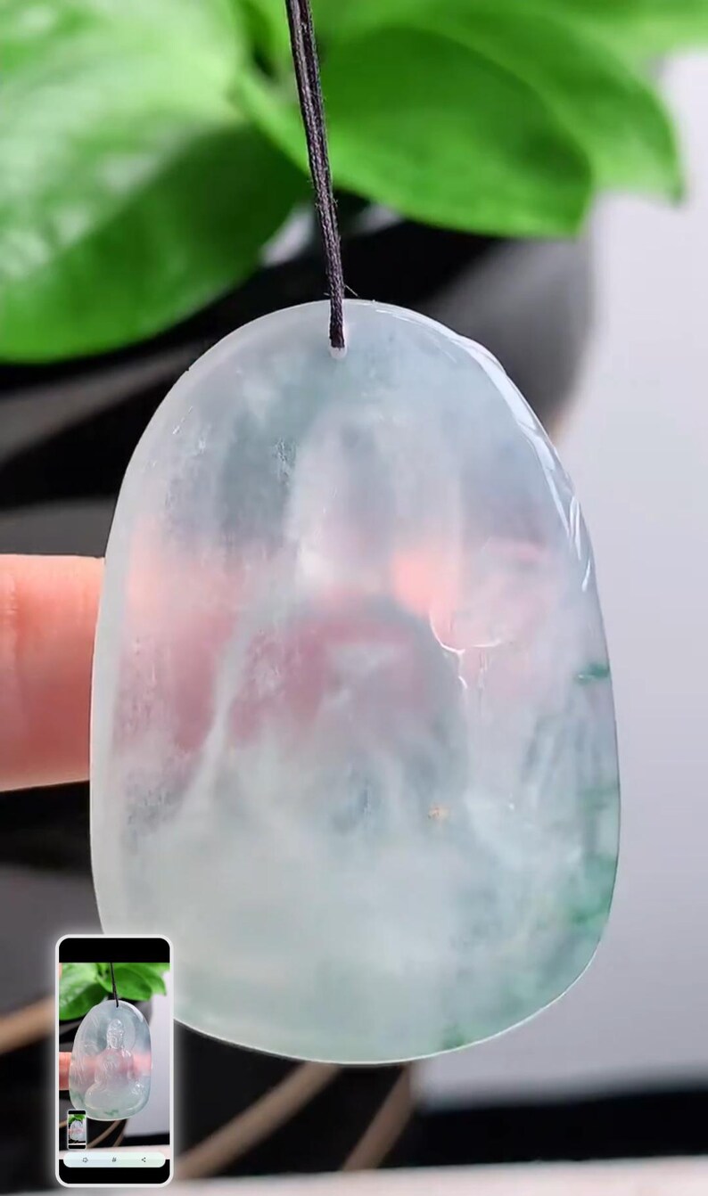 May include: A translucent jade pendant with a light green and pink hue, suspended by a black cord. The smooth, polished surface reflects light, showcasing the natural variations within the stone. The pendant's shape is an elongated oval.